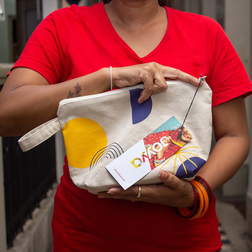 A woman wearing a red fair trade organic v=neck t-shirt holding a cotton canvas pouch between her left and right forearms. The pouch has a blue, black and yellow geometric print and a product tag reading JOYYA hanging from it.