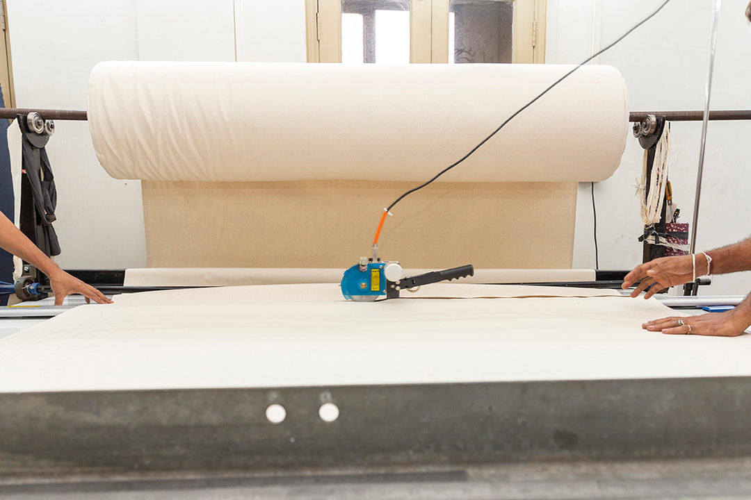A large industrial sized roll of cotton canvas suspended above a cutting table with a fabric cutter going from right to left across the image.