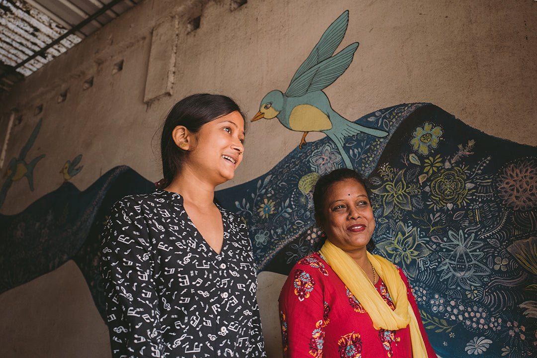 Two Joyya staff women walking in a building front entrance with a mural of a blue bird and blue painted sari fabric on the wall behind them.