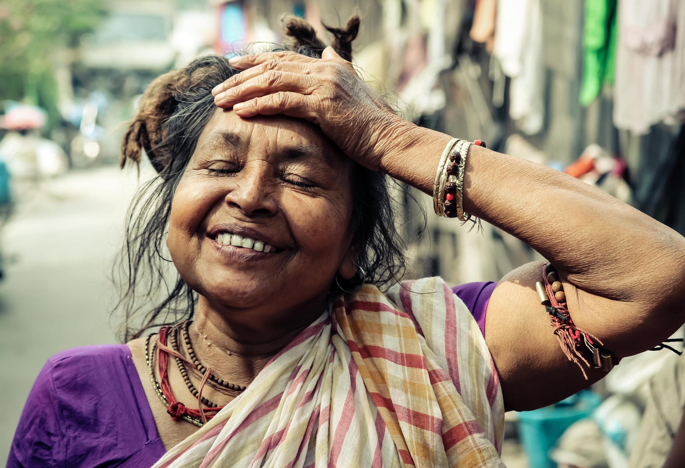 An Indian woman wearing a sari and beads around her arms and neck standing in the streets of an India city with her hand on her forehead while smiling.