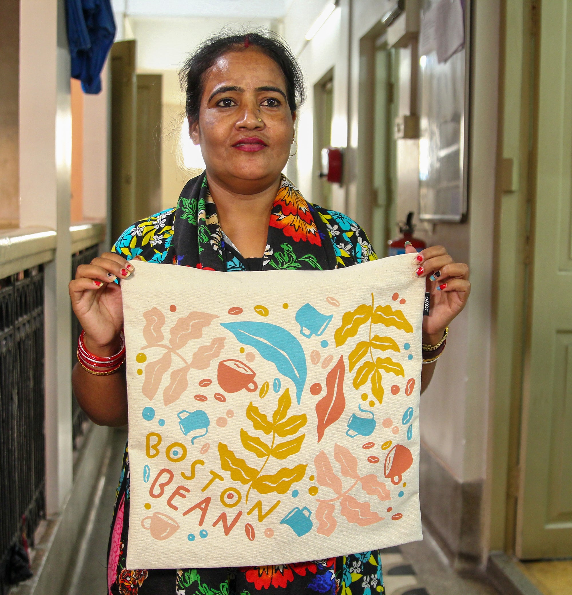 A Joyya staff woman is holding a blank natural color cotton canvas tote bag from each corner showing it to the camera. The bag is printed with a blue, pink, yellow and coral colored design of coffee bean and coffee leaves for the Boston Bean Coffee Company.