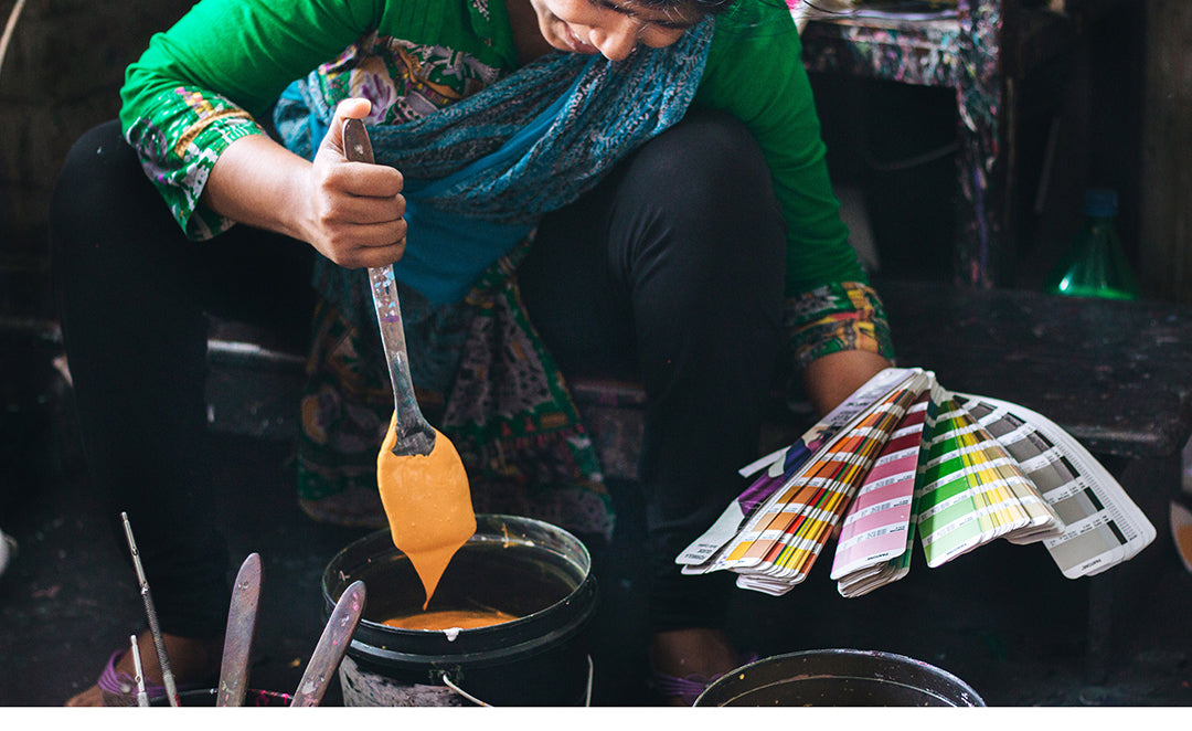 A Joyya Staff woman mixed paint for silk screening a design onto a product. In her right handle is a spatula being used to mix a bucket of yellow paint. In her left hand is a deck of paint chips that she is using to match the paint color.