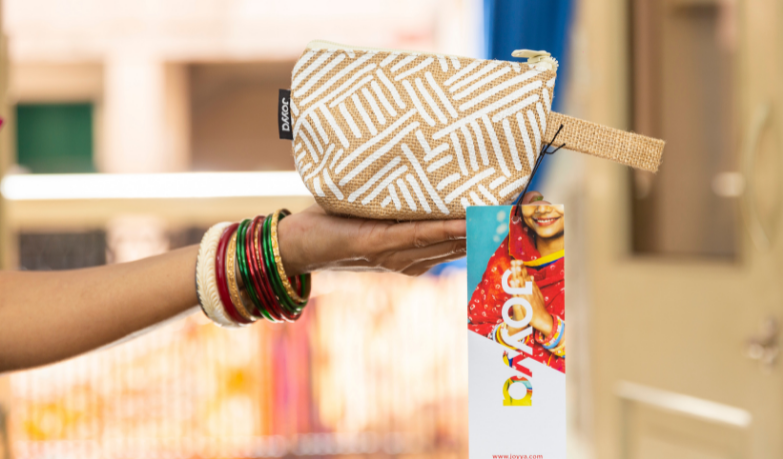 A hand holding up a zippered pouch made from jute printed with a white line geometric print. The bag has a tag hanging from it that reads JOYYA showcasing the manufacturer. 