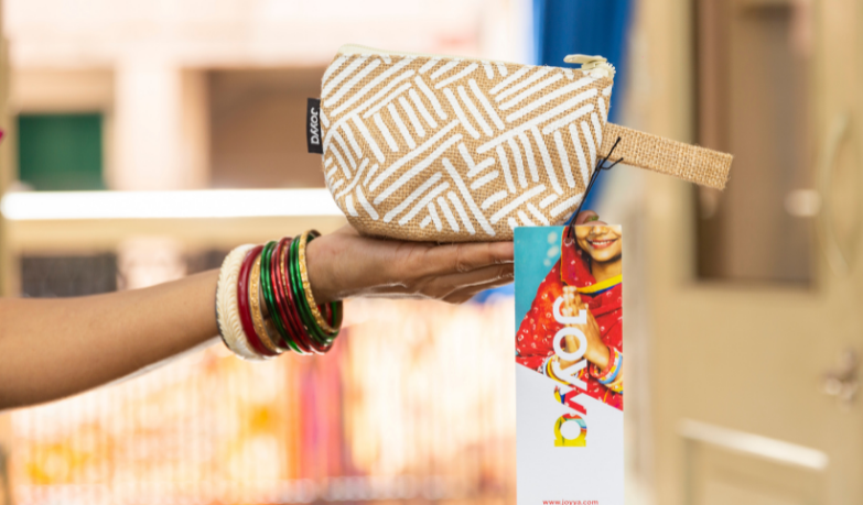 A hand holding up a zippered pouch made from jute printed with a white line geometric print. The bag has a tag hanging from it that reads JOYYA showcasing the manufacturer.