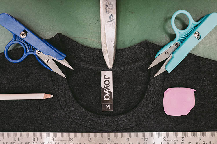 A t-shirt neckline laying flat on a green table top showing the manufacturer label reading JOYYA. Surrounding the next line in a half circle layout are the tools used to fabricate the shirt including fabric markers, scissors, rules and chalk.