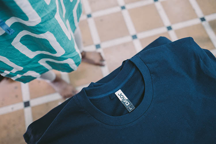 Looking from above down over a table filled with Navy colored organic cotton fair trade t-shirts pilled up for packing. The t-shirt tag on the neckline displays the name of the manufacturer and reads JOYYA.