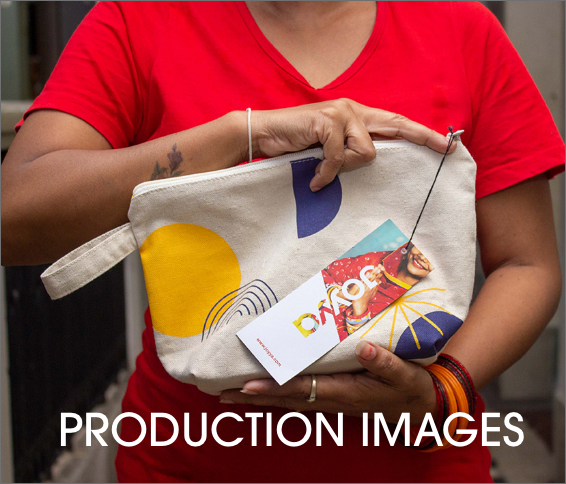 A woman wearing a red fair trade organic v=neck t-shirt holding a cotton canvas pouch between her left and right forearms. The pouch has a blue, black and yellow geometric print and a product tag reading JOYYA hanging from it.