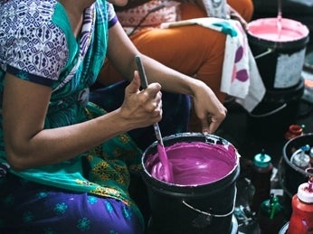 A Joyya Staff woman mixed paint for silk screening a design onto a product. In her right handle is a spatula being used to mix a bucket of purple paint.
