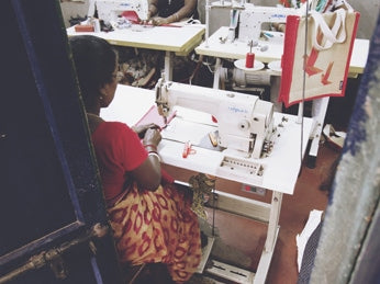 Looking over the shoulder of a women wearing a red and yellow sari sitting at a sewing table with a sewing machine.