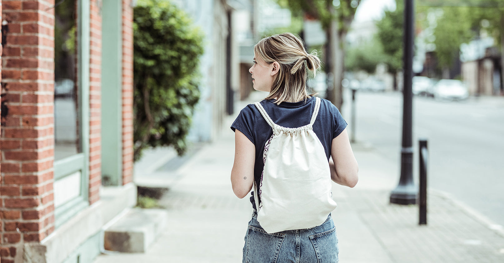 A woman wearing a natural colored cotton canvas backpack with rope drawstrings facing away from the camera.
