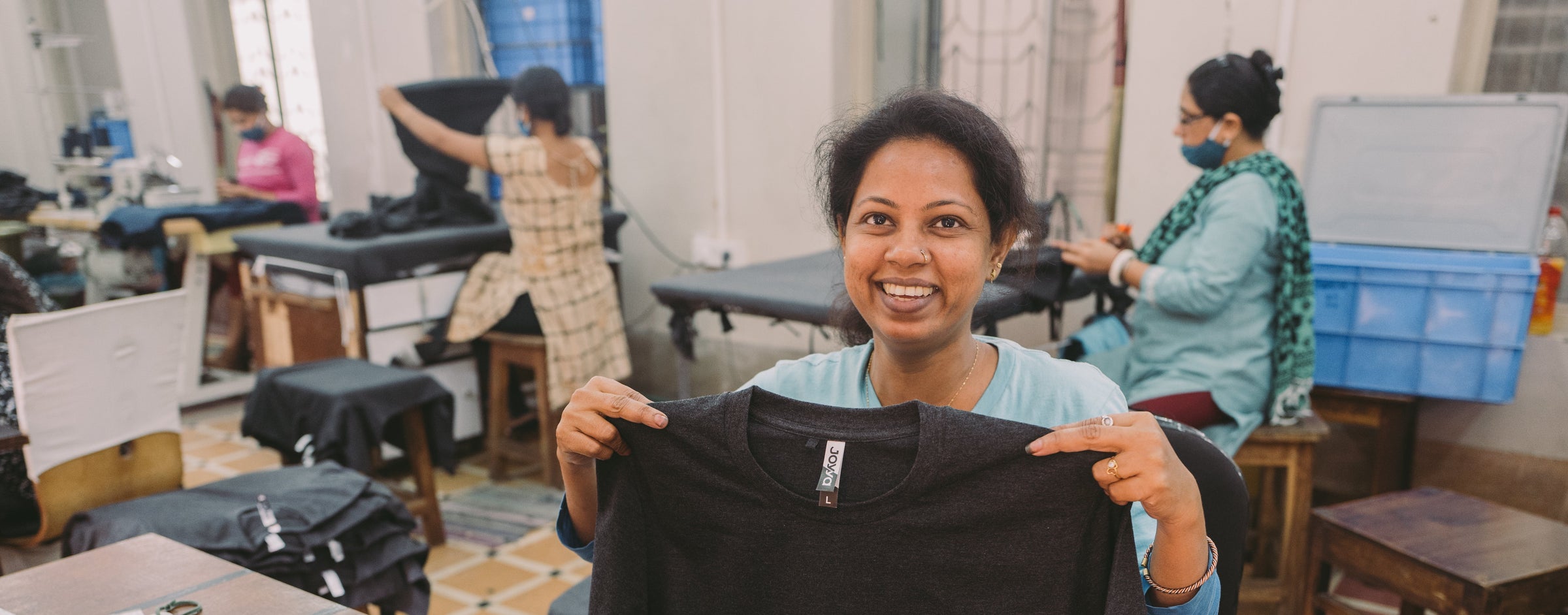 Person holding a black t-shirt in a workshop setting with other people and equipment in the background.