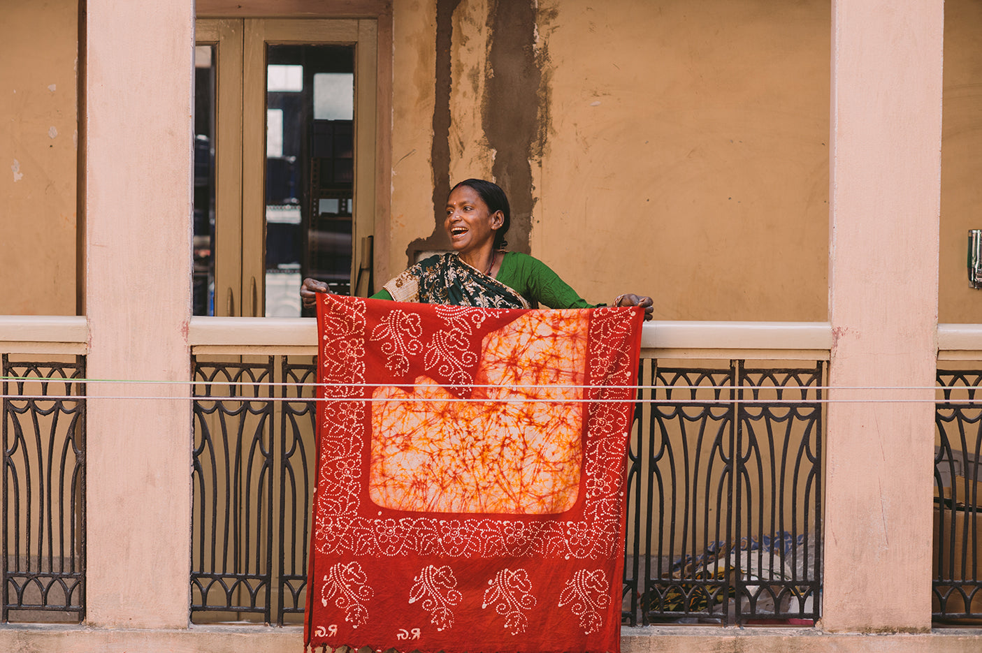 A smiling woman standing on a courtyard balcony hanging a sari on a washing line to dry. 