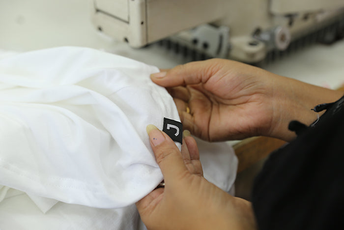 Two hands holding the seam of a white fair trade organic Joyya t-shirt that has just been sown. The hem has a black fabric label with a large letter J for the brand Joyya on it. 