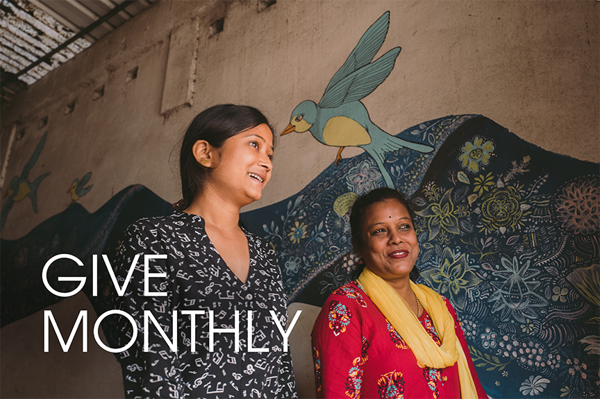Two Joyya staff women walking in a building front entrance with a mural of a blue bird and blue painted sari fabric on the wall behind them.