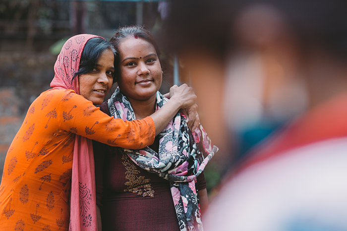 Two staff women of Joyya standing side by side, one with their arms around the other hugging from the side. They are smiling at the camera. 