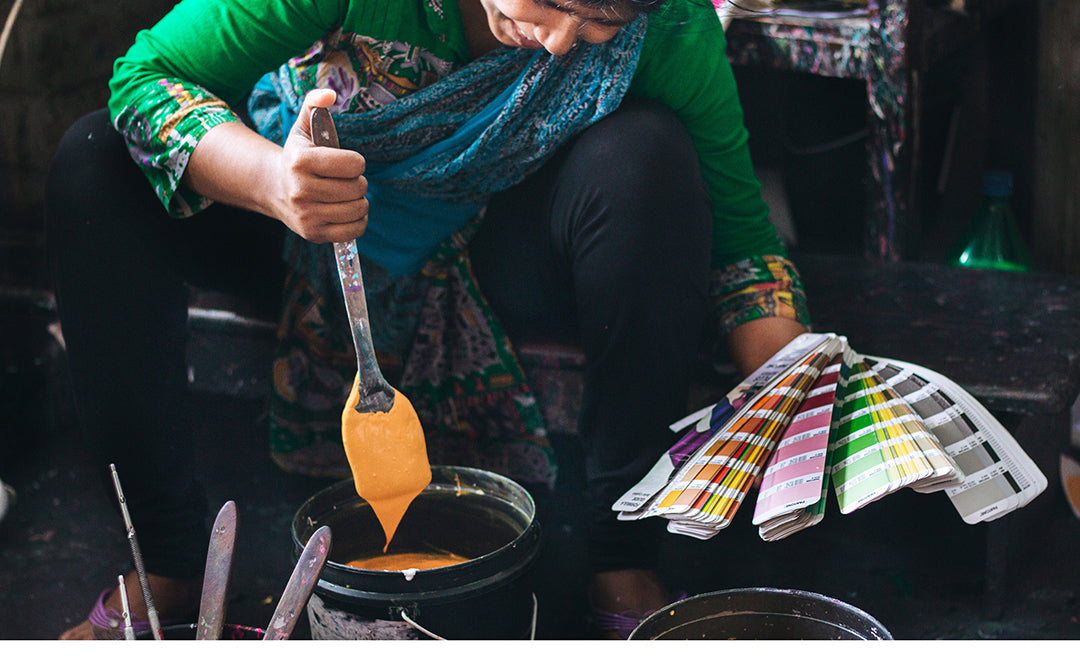 A Joyya Staff woman mixed paint for silk screening a design onto a product. In her right handle is a spatula being used to mix a bucket of yellow paint. In her left hand is a deck of paint chips that she is using to match the paint color. 
