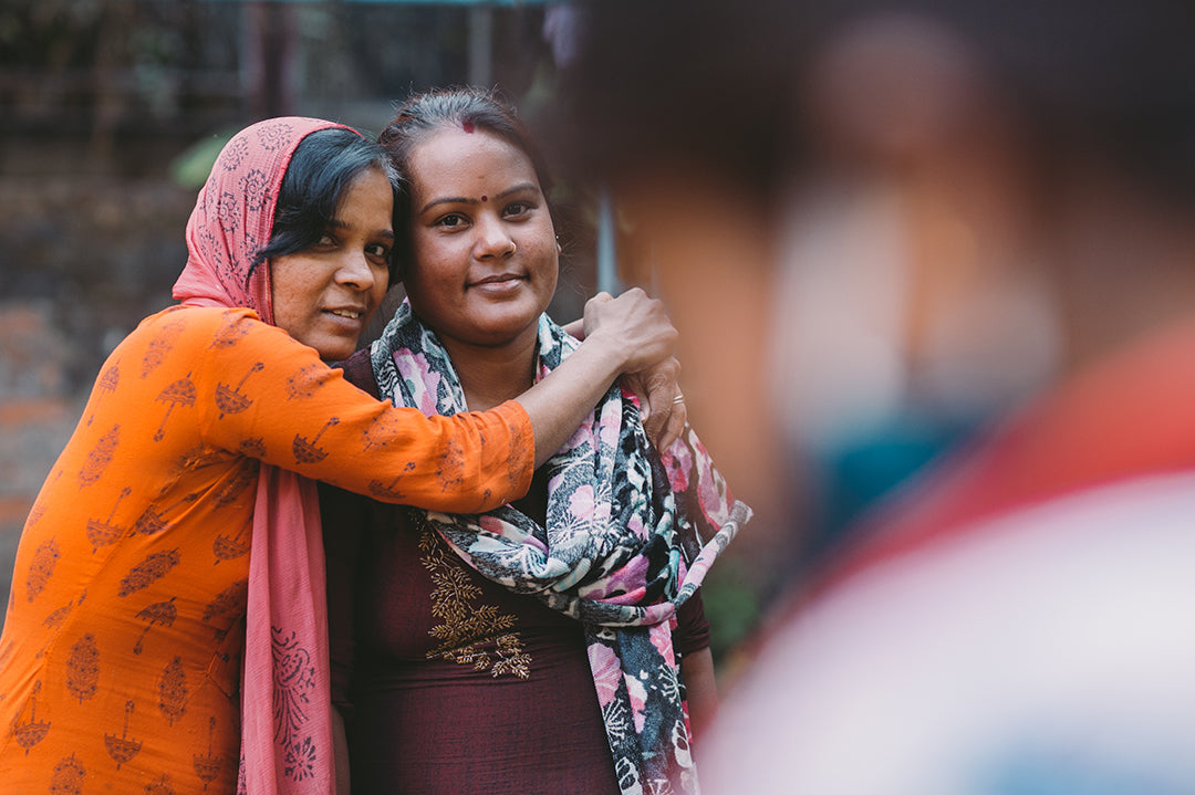 Two staff women of Joyya standing side by side, one with their arms around the other hugging from the side. They are smiling at the camera.