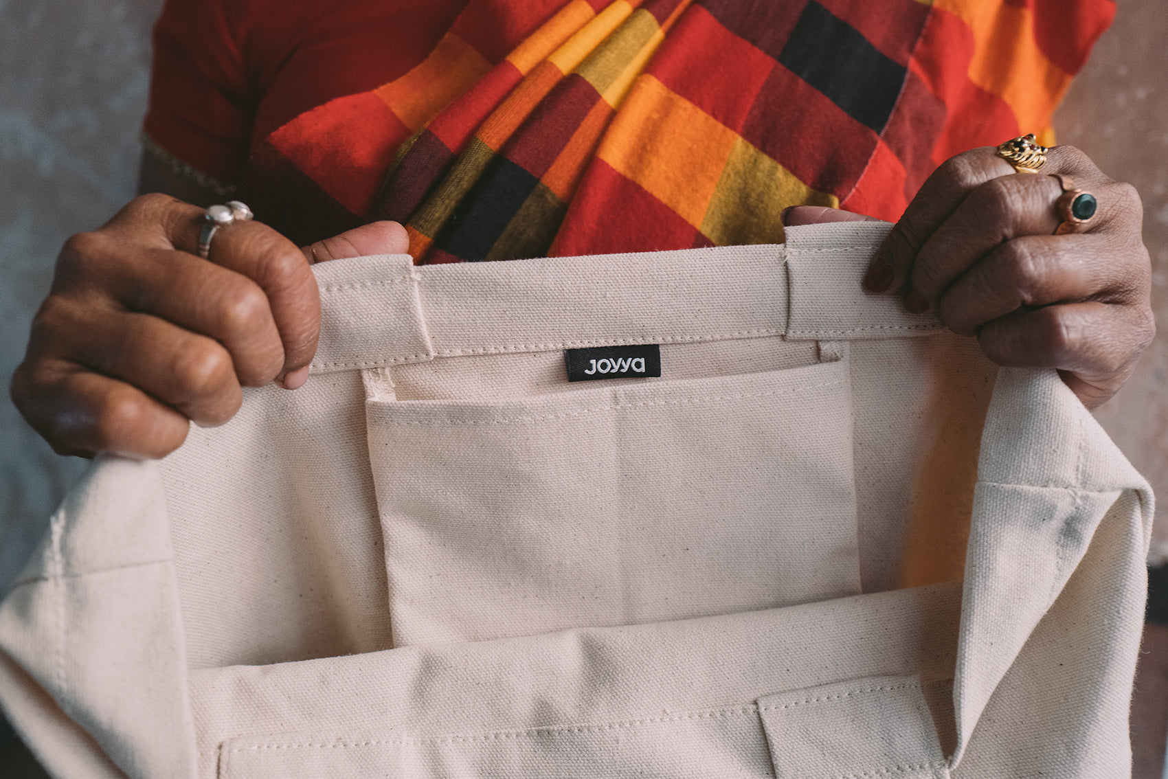 A joyya staff woman holding a natural colored cotton canvas tote bag showing the inside of the bag with a pocket and Joyya fabric label. 