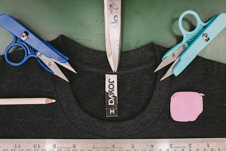 A t-shirt neckline laying flat on a green table top showing the manufacturer label reading JOYYA. Surrounding the next line in a half circle layout are the tools used to fabricate the shirt including fabric markers, scissors, rules and chalk. 