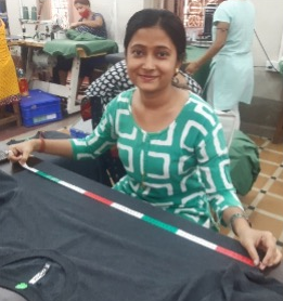 A woman in front of a sewing table with a freshly sewn t-shirt in front of her. She is holding a measuring tape from end to end on the body of the shirt. 