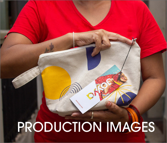 A woman wearing a red fair trade organic v=neck t-shirt holding a cotton canvas pouch between her left and right forearms. The pouch has a blue, black and yellow geometric print and a product tag reading JOYYA hanging from it. 